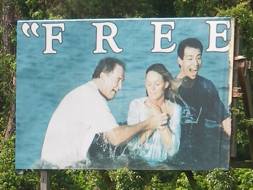 Billboard of three people in a river. Two are getting baptized. Originally, the billboard said "FREEDOM" but half blew down. Now it only says, "Free